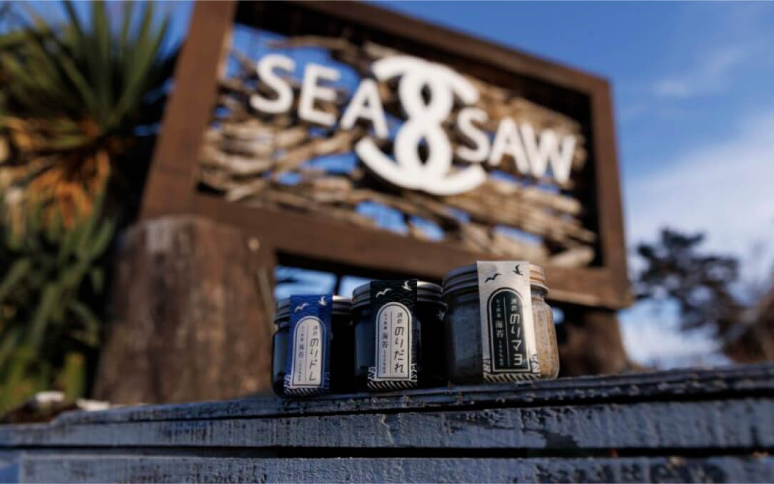 コーヒー缶を前景に置いた「SEA SAW」の看板と店舗ロゴが写る店頭の様子